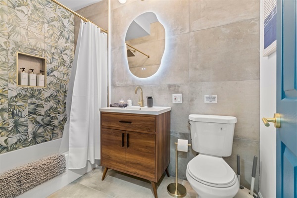 Indulge in a spa-like experience in this elegant bathroom featuring stylish gold accents, a chic backlit mirror, and a beautifully tiled shower. Enjoy modern amenities and a serene atmosphere