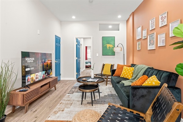 Step into a bright and stylish living room featuring cozy seating, vibrant cushions, and unique decor. The space is enhanced by modern art, lush plants, and a striking gold chandelier, creatin