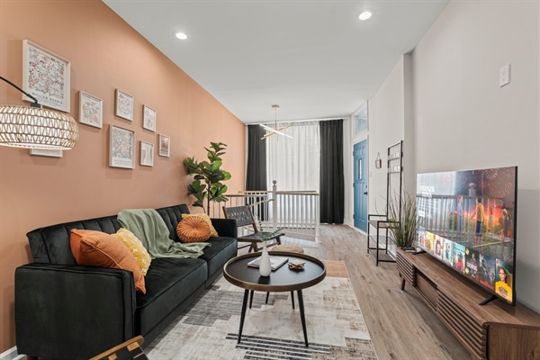 Step into a bright and stylish living room featuring cozy seating, vibrant cushions, and unique decor. The space is enhanced by modern art, lush plants, and a striking gold chandelier, creatin